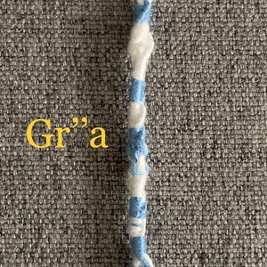 Peut inclure: Un pompon fait à la main avec une boucle en haut, composé de fil blanc et bleu clair torsadé. Le pompon a une frange blanche en bas. Le mot "Gr"a" est écrit en jaune.