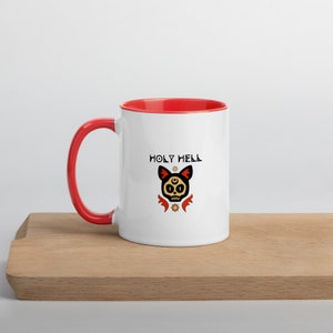 May include: White ceramic mug with a red interior and handle. The mug features the text "HOLY HELL" above a stylized cat skull design with a third eye and star accents. The mug sits on a wooden surface.