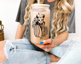 May include: A clear glass tumbler with a black and white bat design and floral accents. The tumbler has a lid and a straw.