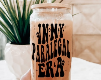 May include: A clear glass jar with a bamboo lid and a straw, filled with a light brown liquid. The jar has a black graphic that says "In My Paralegal Era" with stars and flowers.