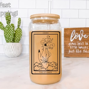 May include: A clear glass tumbler with a bamboo lid and straw, filled with a light brown liquid. The tumbler features a black and tan graphic of a hand holding a cannabis leaf, with the text "THE STONER". A cactus and a wooden sign are in the background.
