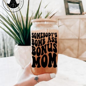 Op de afbeelding: Een herbruikbare drankbeker van helder glas met een bamboe deksel en een rietje. De beker heeft een zwarte vinyl sticker met de tekst "Somebody's Bomb Ass Bonus Mom" in een retro lettertype.