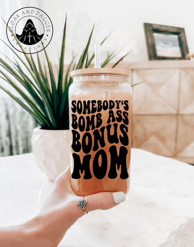 Op de afbeelding: Een herbruikbare drankbeker van helder glas met een bamboe deksel en een rietje. De beker heeft een zwarte vinyl sticker met de tekst "Somebody's Bomb Ass Bonus Mom" in een retro lettertype.