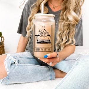 May include: A clear glass tumbler with a brown lid and a black and white illustration of a typewriter with flowers growing out of it. The tumbler has the text "XII THE WRITER." printed on it.