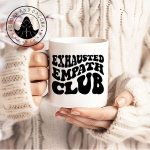 May include: White ceramic mug with a black graphic that reads "EXHAUSTED EMPATH CLUB" in a retro, wavy font.