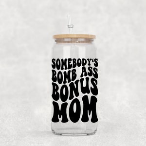 Op de afbeelding: Een doorzichtige glazen beker met een houten deksel en een rietje. De beker heeft een zwarte vinyl sticker met de tekst "Somebody's Bomb Ass Bonus Mom" in een retro-lettertype.