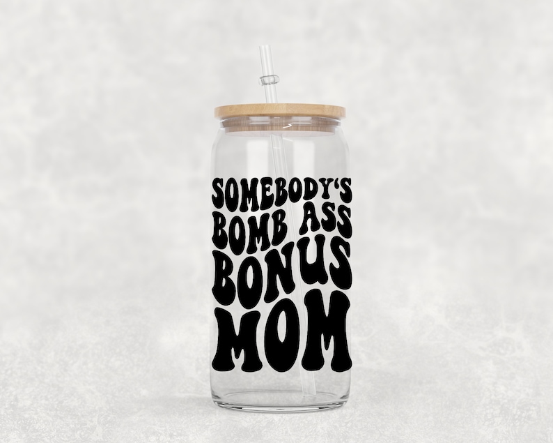 Op de afbeelding: Een doorzichtige glazen beker met een houten deksel en een rietje. De beker heeft een zwarte vinyl sticker met de tekst "Somebody's Bomb Ass Bonus Mom" in een retro-lettertype.