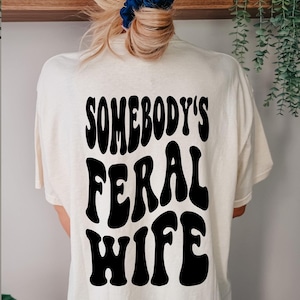 Puede incluir: Una camiseta blanca con el texto "Somebody's Feral Wife" impreso en negro, en una fuente de estilo retro.