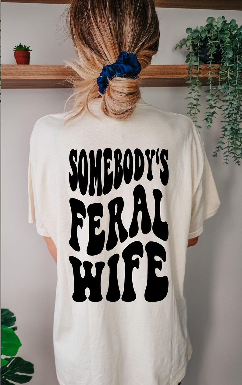 Puede incluir: Una camiseta blanca con el texto "Somebody's Feral Wife" impreso en negro, en una fuente de estilo retro.