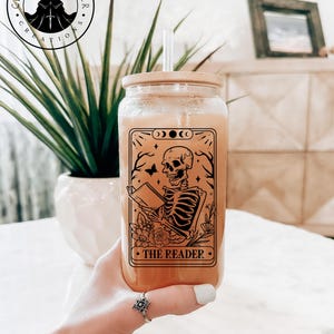 May include: A clear glass tumbler with a bamboo lid and straw. The tumbler features a tarot card design with a skeleton reading a book, the text "THE READER", and the "Cloak and Dagger Creations" logo.