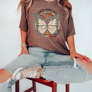 May include: A brown t-shirt with a graphic of a butterfly with the text "Anti-Social Butterflies" printed on it. The person is wearing blue jeans and brown sneakers.