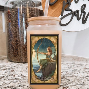 May include: A clear glass tumbler with a brown wooden lid and a straw. The tumbler has a graphic of a woman with a bow and arrow, with the text "SAGITTARIUS" below the image.