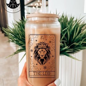 May include: A glass jar with a wooden lid and a straw, filled with a brown liquid. The jar has a black and white design of a lion with a crown, stars, and clouds. The design includes the text "The Leo" and the astrological symbol for Leo.