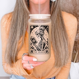 May include: A clear reusable tumbler with a white lid and a straw. The tumbler features a black and white illustration of a skeleton reading a book with the text "THE READER" below the image.