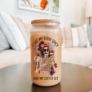 May include: A glass tumbler with a bamboo lid and a straw. The tumbler has a graphic of a skeleton holding a book and a coffee cup. The text on the tumbler reads "I like my book spicy and my coffee icy."