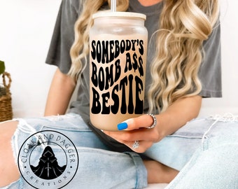 May include: A clear reusable tumbler with a white lid and a black graphic that says "Somebody's Bomb Ass Bestie".