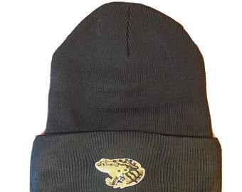 May include: Black beanie with a folded brim. The beanie features a small embroidered patch of a frog in shades of green, tan, and white. The hat is made of a soft, knitted material.