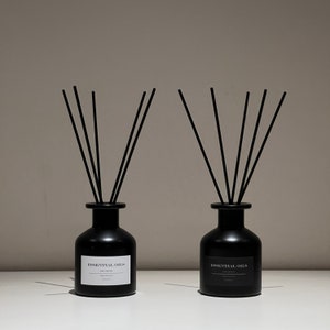 May include: Two black essential oil diffusers with black reeds. Each diffuser has a matte black bottle with a white label that reads "ESSENTIAL OILS". The bottles are on a white surface against a neutral background. The reeds are arranged in a fan shape.