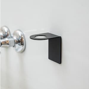 Puede incluir: Un portacepillos de dientes de metal negro montado en una pared blanca. El soporte tiene una abertura circular para un cepillo de dientes y una base rectangular.