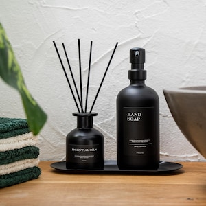 Könnte beinhalten: Schwarzer ätherischer Öldiffusor mit schwarzen Schilfrohren und eine schwarze Handseifenflasche mit Pumpspender. Beide Flaschen haben weiße Etiketten mit schwarzem Text. Die Flaschen stehen auf einem schwarzen Tablett.