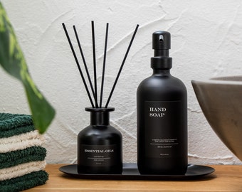 May include: Black essential oil diffuser with black reeds and a black hand soap bottle with a pump dispenser. Both bottles have white labels with black text. The bottles are on a black tray.