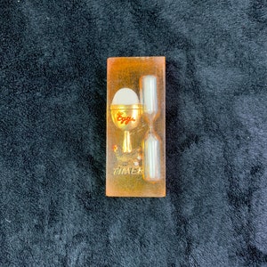 May include: A clear plastic egg timer with a gold-colored base and a white egg inside. The timer has a red inscription that says "Egg Timer".