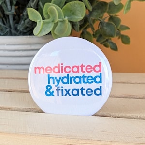 May include: White button with the text "medicated hydrated & fixated" in pink, blue, and dark blue lettering.