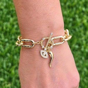 May include: A gold-tone chain link bracelet featuring a toggle clasp closure. The bracelet includes two dangling charms: one shaped like an eye with embedded clear stones and one curved horn-shaped charm. The toggle bar is also accented with clear stones.
