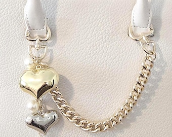 May include: A white handbag with a textured finish, featuring a gold chain and heart-shaped charms. The bag has two handles and a zipper closure. The charms include a gold heart, a silver heart, and faux pearls.