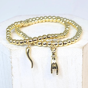 May include: A set of two gold-toned beaded stretch bracelets. One bracelet features a small, curved horn-shaped charm, while the other bracelet displays a hand-shaped charm with the index and pinky fingers extended in a gesture.
