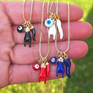 May include: Gold-tone necklaces with various charms, including a black hand, a white hand, a red hand, and a blue hand. Each charm is paired with an evil eye pendant and a horn-shaped charm. The necklaces are displayed against a green background.