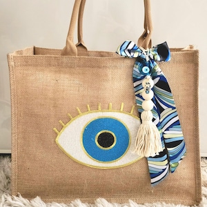 May include: A brown jute tote bag with a large blue evil eye design. The bag has a white tassel and a blue and white patterned scarf attached to the handle.