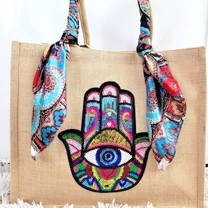 May include: A beige burlap tote bag with a colorful Hamsa hand design. The bag features a decorative scarf tied to the handles with a paisley pattern in shades of blue, red, and orange. The Hamsa hand is embroidered with sequins.