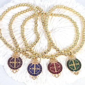 May include: Four gold beaded bracelets with colorful Saint Benedict medals. Each medal features a cross and text in a circular design, with colors including purple, blue, red, and green. The bracelets are displayed on a white, textured surface.