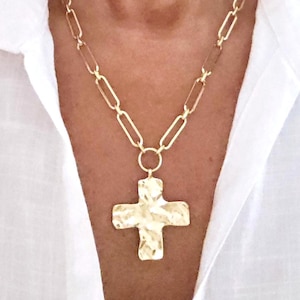May include: Gold-toned necklace featuring a large, textured cross pendant. The necklace has a chain of elongated links and a smaller ring connecting the cross. The cross has a hammered appearance. The necklace is worn over a white collared shirt.
