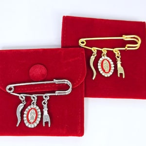 May include: Two safety pin brooches, one silver-toned and one gold-toned, each featuring three dangling charms: a curved horn, an oval medallion with a red center and clear rhinestones, and a hand gesture charm, resting on red velvet pouches.