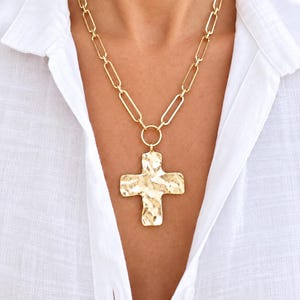 May include: A gold-toned necklace featuring a long paperclip chain and a large, central cross pendant. The cross has a hammered, textured surface finish and is attached to the chain by a circular jump ring, resting against a white collared shirt.