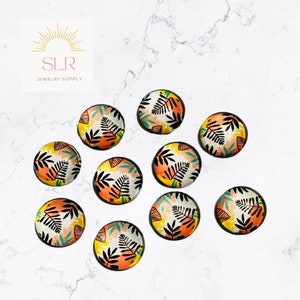 May include: Ten round cabochons with a tropical leaf design. The cabochons have a white background with black, orange, yellow, and teal leaf patterns. The SLR Jewelry Supply logo is in the upper left corner.