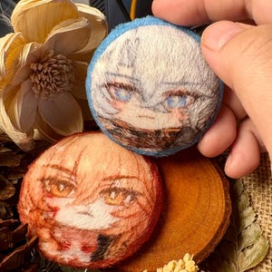 以下が含まれることがあります： 2つのぬいぐるみ、丸いキャラクターピン。1つのピンは青い縁取りと青い目の白髪のキャラクターです。もう1つのピンは、オレンジ色の髪のキャラクターとオレンジ色の目です。どちらのピンにも詳細な顔の特徴があります。