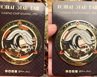May include: Gold-coloured enamel pin featuring a character with white hair and red eyes. The character is wearing a black and red outfit and is surrounded by a gold and red circle with the text "HONKAI STAR RAIL CASINO CHIP ENAMEL PIN".