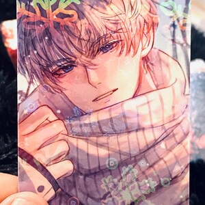 May include: A rectangular card with an anime-style illustration of a person with light hair and blue eyes. The person is wearing a scarf and surrounded by snowflake designs. The artwork has a soft, pastel colour palette.