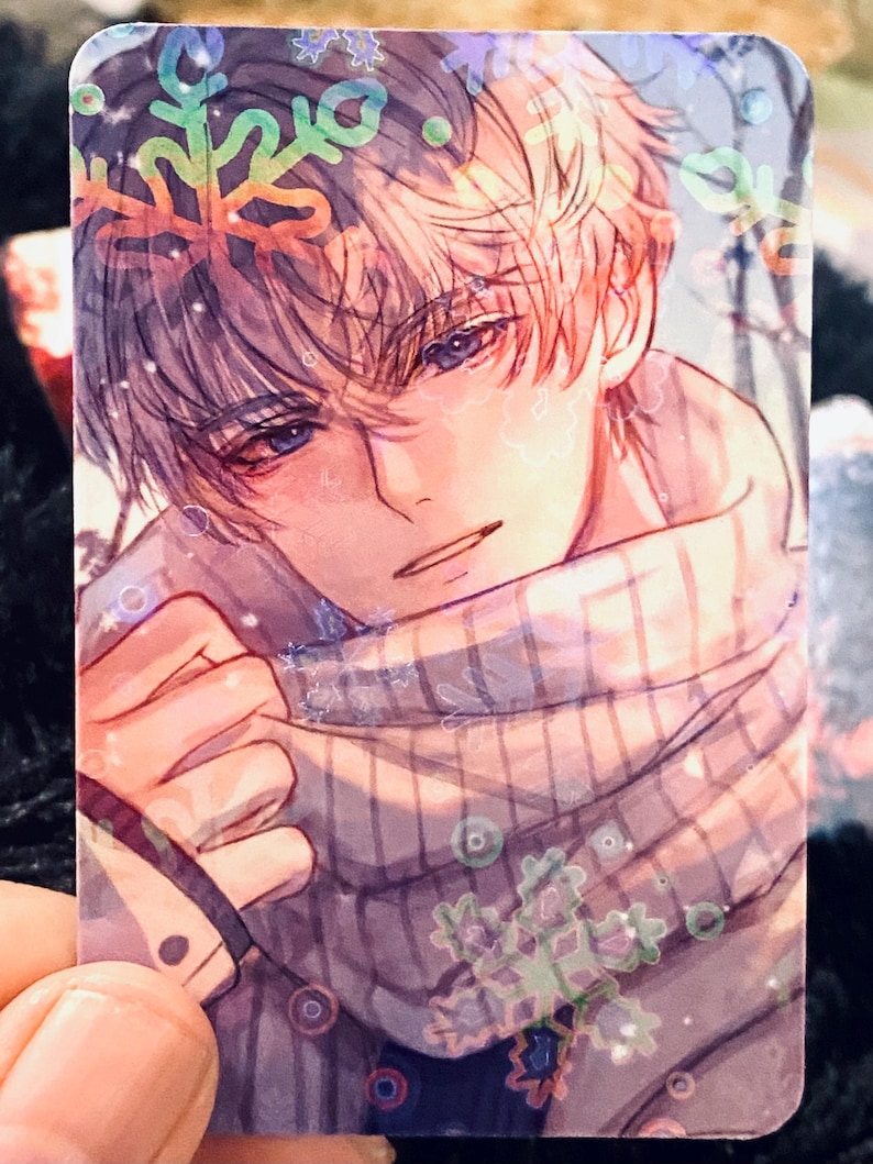 May include: A rectangular card with an anime-style illustration of a person with light hair and blue eyes. The person is wearing a scarf and surrounded by snowflake designs. The artwork has a soft, pastel colour palette.