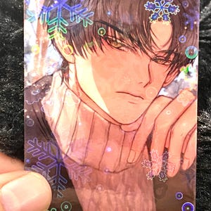 May include: A holographic trading card with an anime-style illustration of a person with dark hair and a sweater. The card features snowflake and bubble accents, with a soft, warm colour palette.