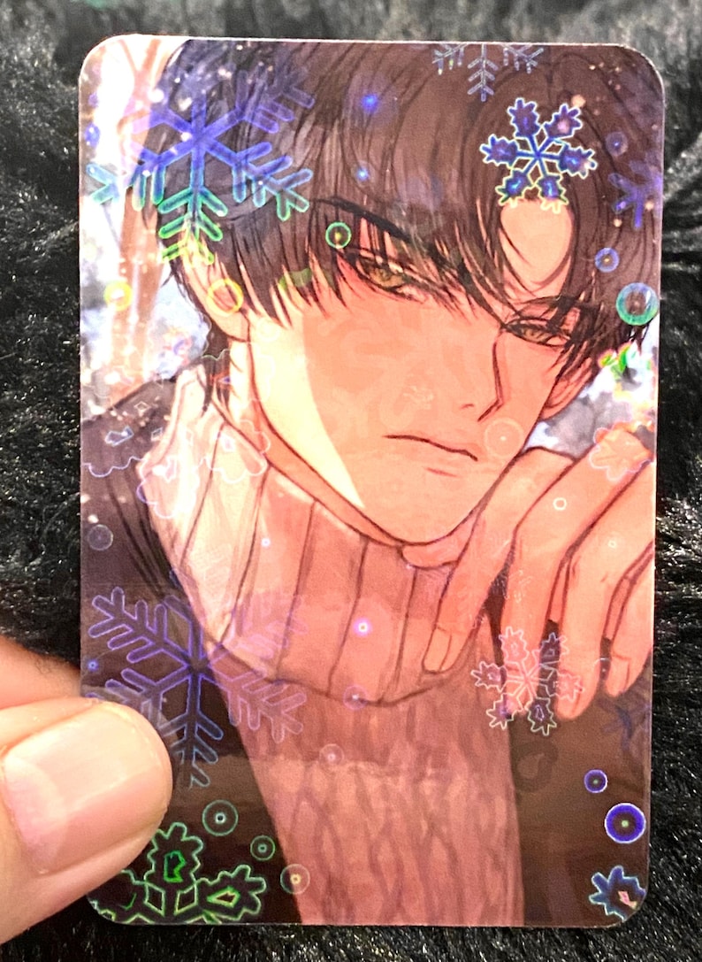 May include: A holographic trading card with an anime-style illustration of a person with dark hair and a sweater. The card features snowflake and bubble accents, with a soft, warm colour palette.