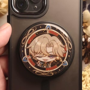 May include: A black phone case with a round, gold-rimmed pop socket featuring a cartoon character with blonde hair, a black eye patch, and a mischievous expression. The character is wearing a gold earring and a black and gold outfit.