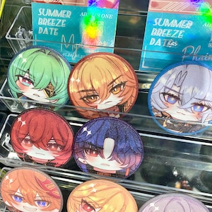 May include: A display of colorful anime-style character button pins. The pins feature various characters with different hair colors, including green, orange, red, blue, and blonde. Each pin has a unique facial expression. The display also includes promotional cards with text.