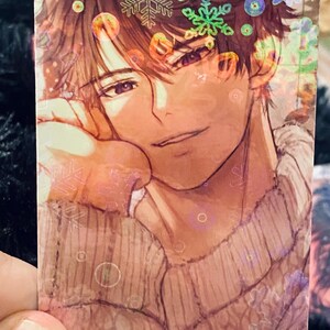 May include: A rectangular card with an anime-style illustration of a person with brown hair and a sweater. The image has a winter theme with snowflake and circular accents. The card has a holographic finish.
