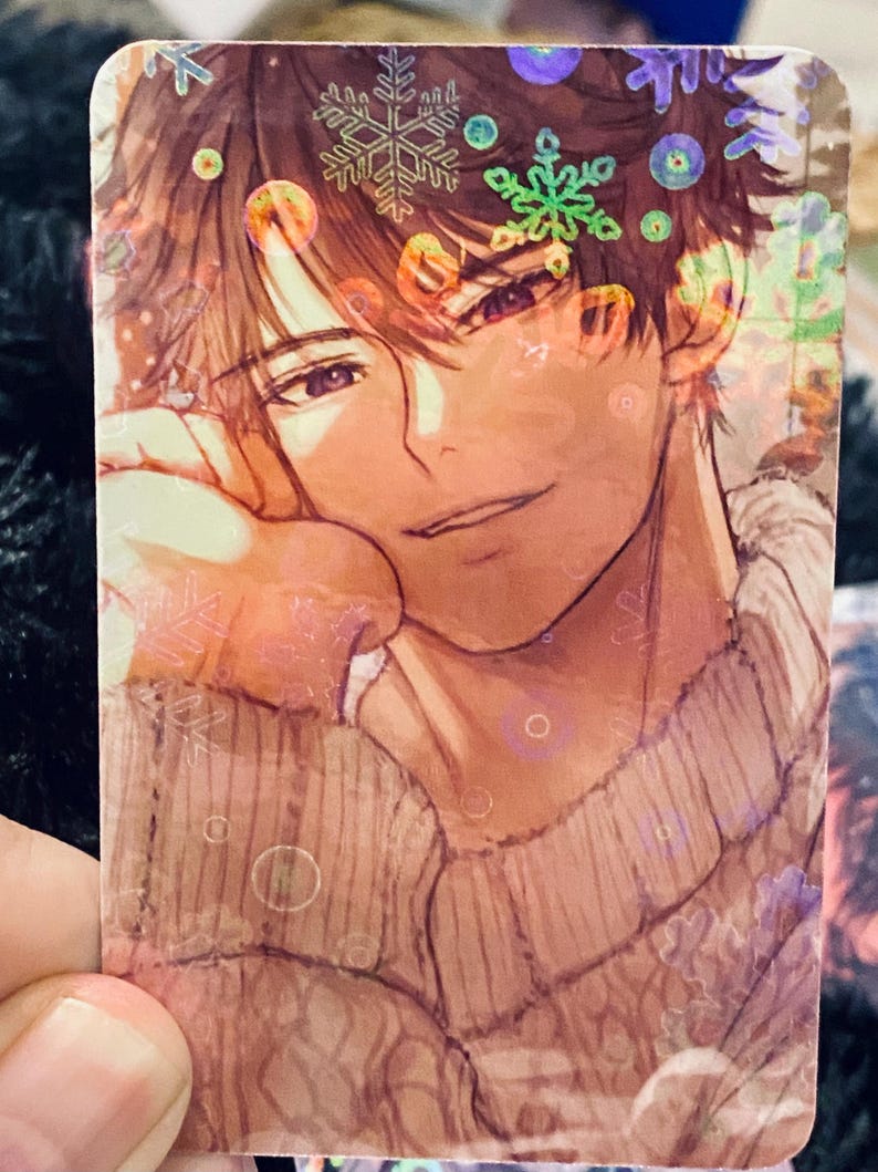 May include: A rectangular card with an anime-style illustration of a person with brown hair and a sweater. The image has a winter theme with snowflake and circular accents. The card has a holographic finish.