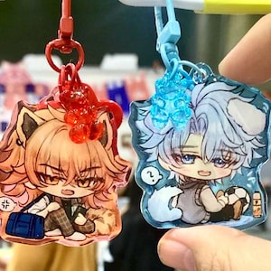 May include: Two anime-style acrylic keychains. One features a character with tan hair, a blue bag, and a red clip. The other has a character with blue hair, a soccer ball, and a blue clip. Both have glitter accents.