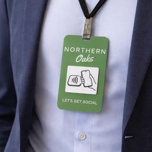 May include: A green rectangular badge with the text "NORTHERN Oaks" and "LET'S GET SOCIAL" is hanging from a black lanyard. The badge features a graphic of a smartphone and a wireless symbol. The person is wearing a blue blazer and a light blue button-down shirt.